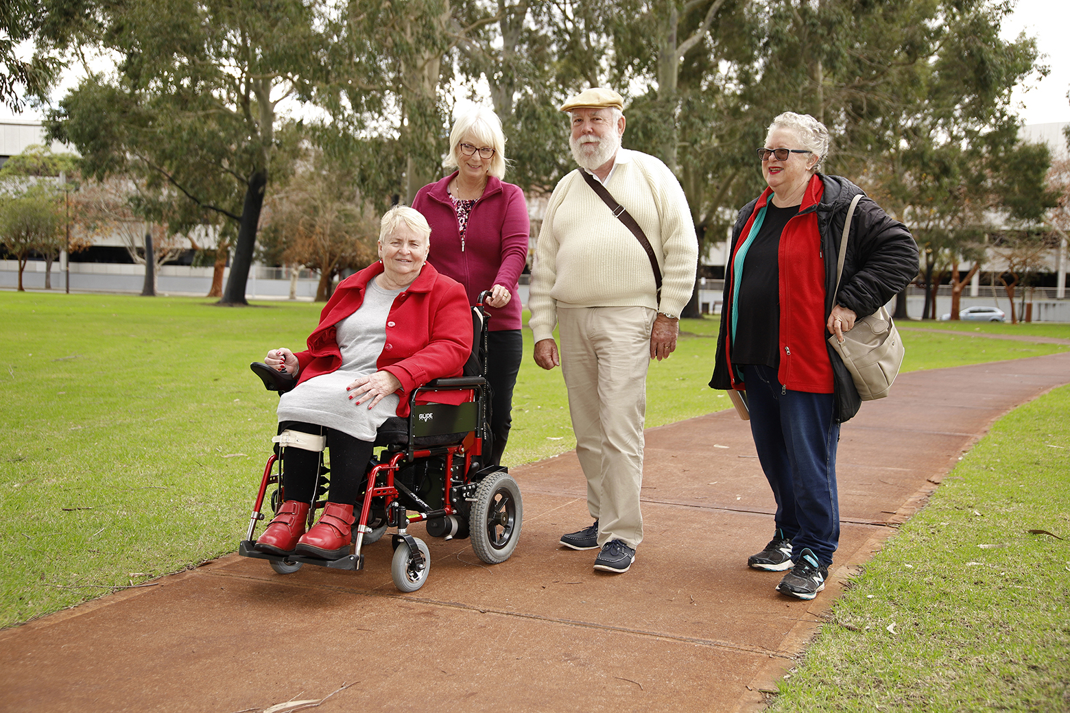 One woman in wheel chair with two older women and one older man standing in a park