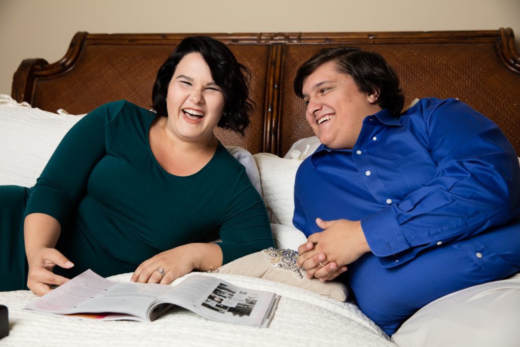 Young man and young women laying on bed reading magazine laughing and smiling at camera
